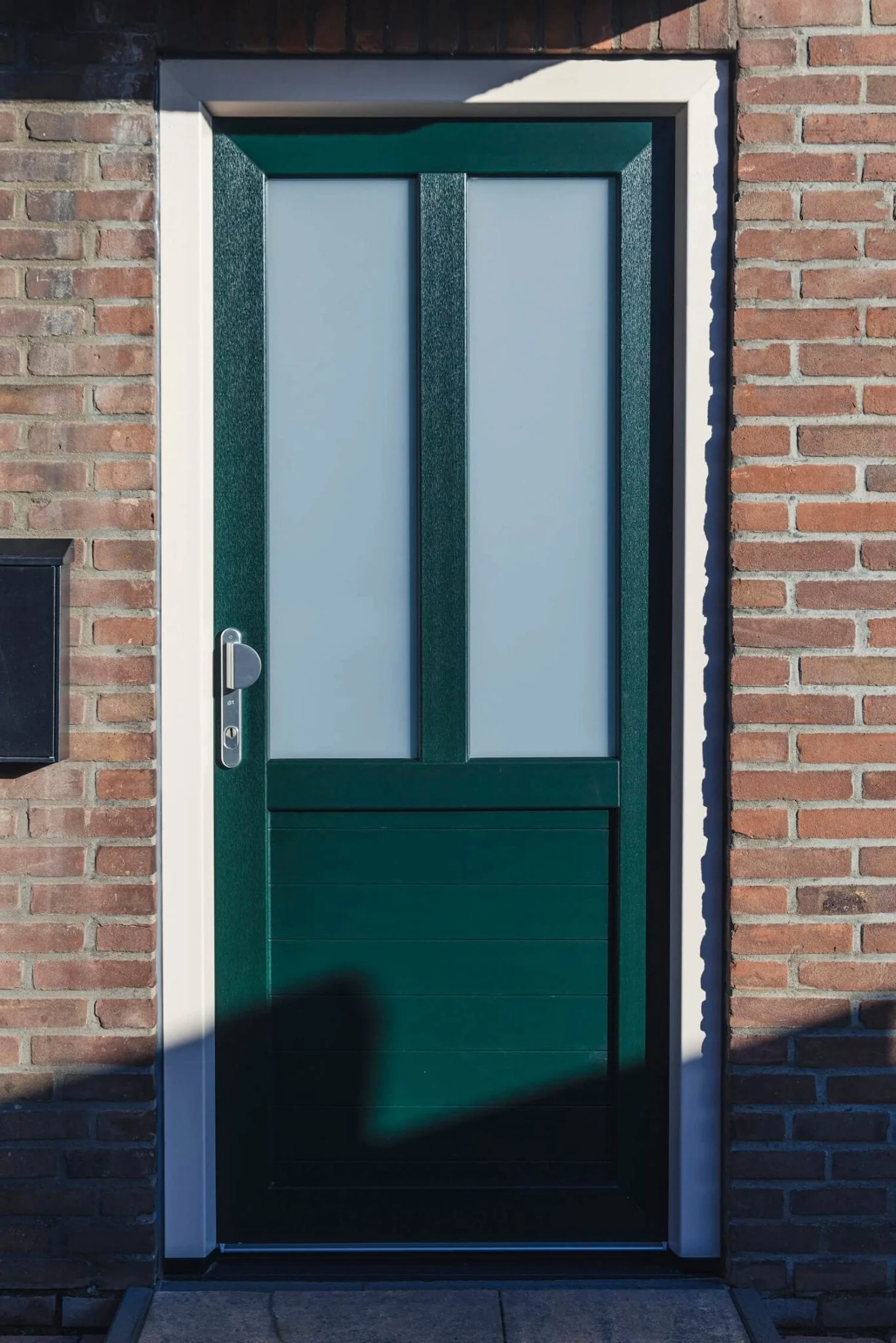 Groene voordeur van kunststof met twee verticale matglazen panelen en een dichte onderzijde, geplaatst in een bakstenen gevel.